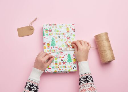 two female hands are wrapping a gift in holiday paper on a pink background, top view, do-it-yourself...