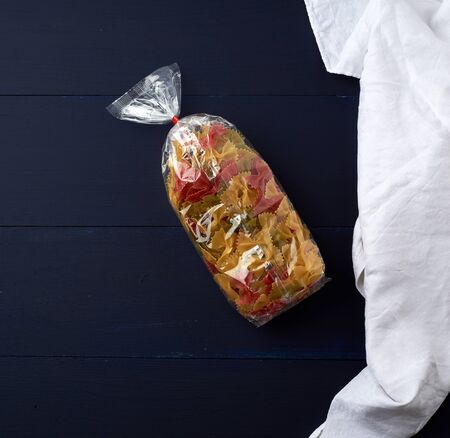 transparent cellophane bag with raw colorful pasta in the form of bows on a blue wooden surface, copy spaceの写真素材