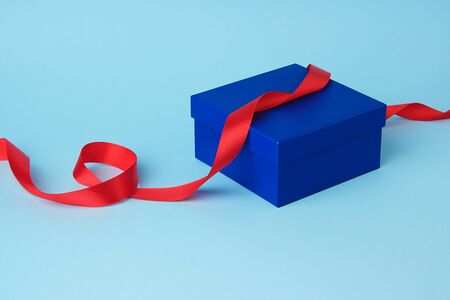 closed square blue cardboard box for a gift and twisted silk red ribbon on a blue background....
