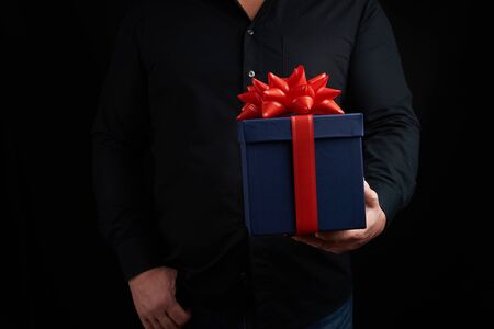 adult man in a black shirt holds a blue square box with a red bow tied on a dark background, concept of congratulations, surpriseの写真素材