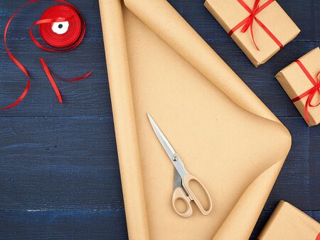 roll of brown kraft packaging paper, box tied with a red silk ribbon, scissors and a bobbin with ribbon on a blue wooden background, top viewの写真素材