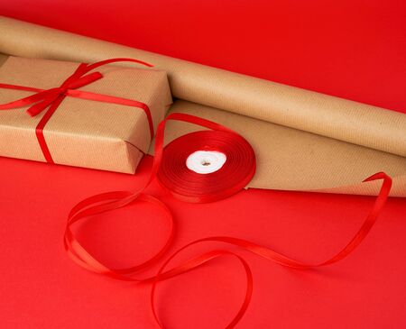 roll of brown paper for packaging, a reel of red ribbon and a wrapped gift in brown kraft paper on a red background, making surprisesの写真素材