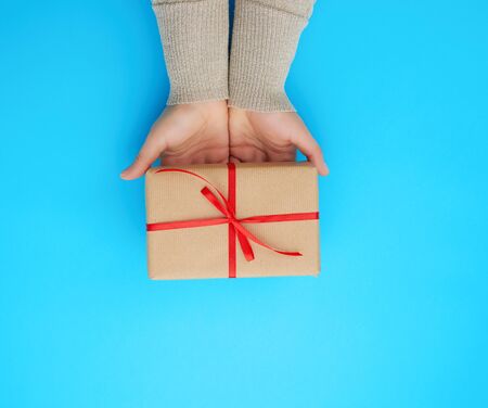 female hand holds a brown box with a red bow on a blue background, concept of a congratulation on a...