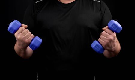 adult male athlete in black uniform holds blue plastic blue dumbbells in hands, black background