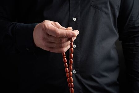 man in a black shirt holds a brown stone rosary in his left hand, low key. used to count prayers or...