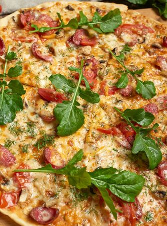baked round pizza with smoked sausages, mushrooms, tomatoes, cheese and arugula leaves, food is cut in portionsの写真素材