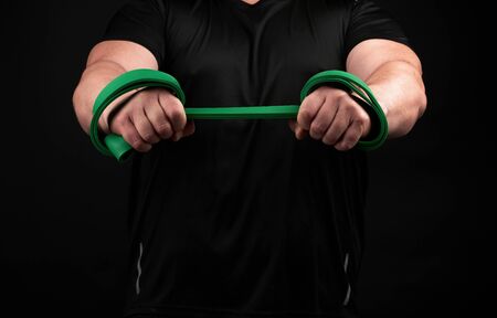 athlete with a muscular body in black clothes is doing physical exercises with green rubber, low key