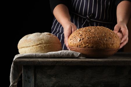 woman in a blue striped apron holds in her hands baked round rye bread and with sunflower seeds, wooden tableの写真素材