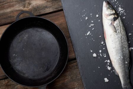 fresh whole sea bass fish on a black board, next to it is an empty round black frying pan on a wooden table, top viewの写真素材
