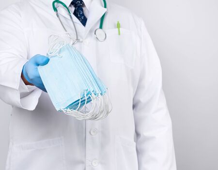 doctor in a white coat, blue latex sterile gloves holds textile medical masks in his hand, protective accessory against viruses and bacteria, close upの写真素材