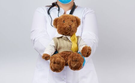 pediatrician in white coat, blue latex gloves holds a brown teddy bear with a yellow ribbon on a sweater, concept of the fight against childhood cancer, problem of suicidesの写真素材