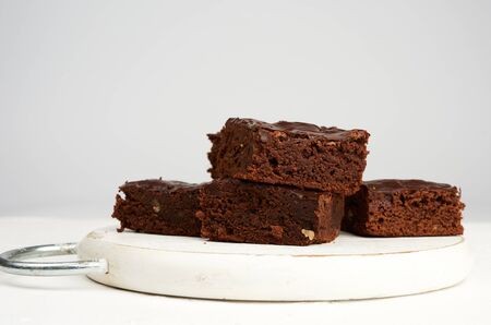 stack of square baked slices of brownie chocolate cake with walnuts on a round white wooden board,...