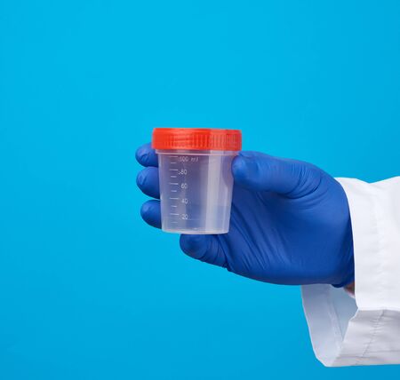 male doctor in a white coat holds a empty plastic container for urine specimen, wearing blue sterile...