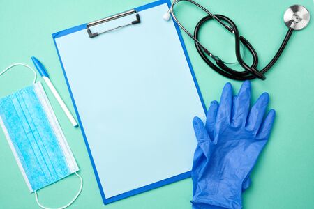 metal stethoscope, paper holder with a blank blue sheet, disposable blue medical mask on a green background, top view, flat layの写真素材
