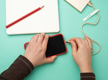 two hands holding a red mobile smartphone and a charging cable on a green background, top view
