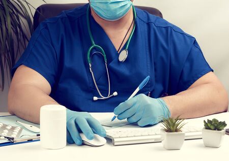 male doctor sitting at a white table in blue uniform and latex gloves, specialist writes a...