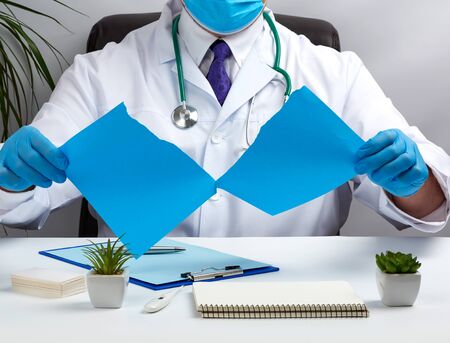 doctor in a white medical coat is sitting at a table in a brown leather chair and holding a torn...