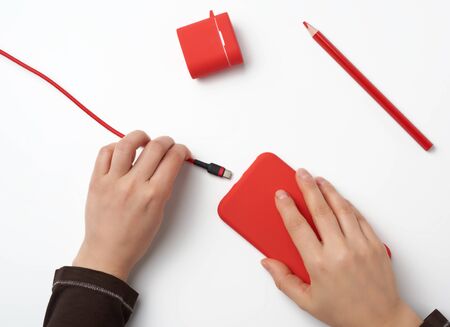 two hands holding a red mobile smartphone and a charging cable on a white background, top viewの写真素材