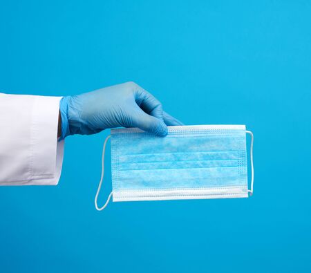 doctor in blue latex sterile gloves holds a blue disposable mask made of non-woven material on a...
