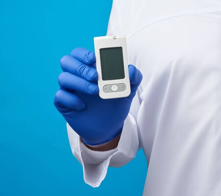male hand with blue sterile gloves holds a white blood glucose meter for measuring blood sugar, blue...