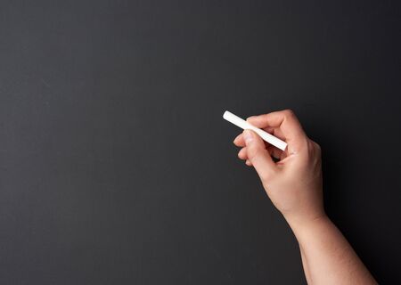 hand holds a piece of white chalk on the background of an empty black chalk board, presentation concept, back to schoolの写真素材