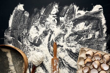sprinkled white wheat flour, wooden rolling pin, round sieve and chicken eggshell in a paper tray,...