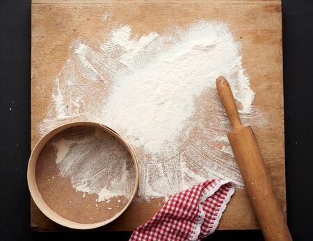 white wheat flour scattered on a black table and a wooden rolling pin, round sieve on a black table,...