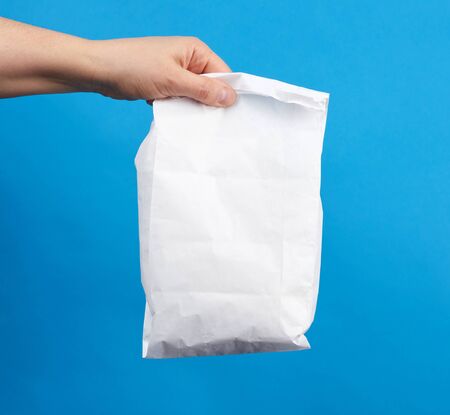 female hand holds a disposable full white paper bag, takeaway and delivery food concept, blue...