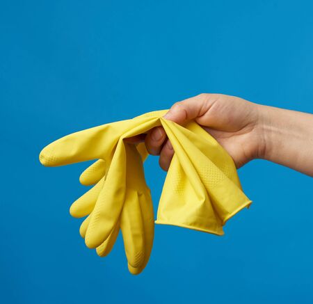 female hand holds yellow  rubber gloves for cleaning, blue background, protective clothing for house...