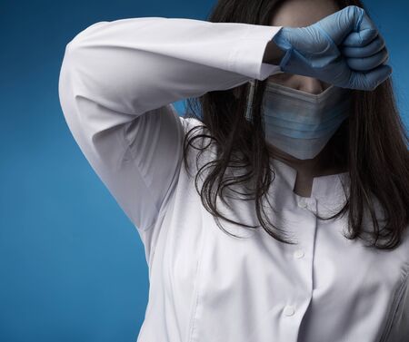 young woman doctor in a white coat, blue latex gloves and a protective mask on his face stands on a...