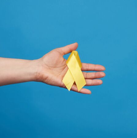 female hand hold a yellow ribbon in the form of a loop on a blue background, symbol of the fight against the cat's tumor, problem of suicides and their prevention, close upの写真素材