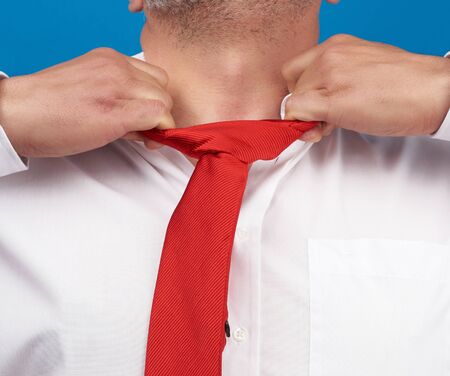 man in a white office shirt tears off a red satin tie from his neck on a blue background, concept of depression, unemployment and dismissalの写真素材