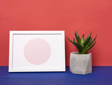 white photo frame and ceramic pot with a growing plant on a blue wooden table, minimalist style, red...