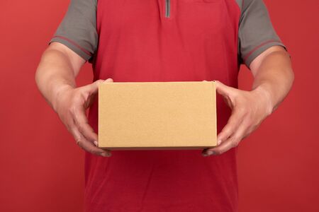 adult man in a red T-shirt holds a cardboard brown box on a red background, concept of delivery of...