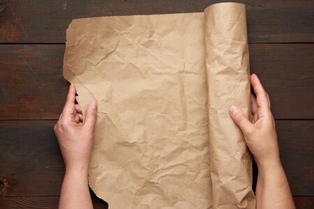 two female hands hold a rolled roll of brown craft paper on a wooden table, top view
