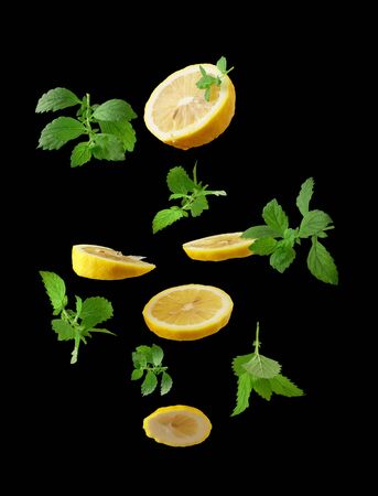 slices of ripe juicy yellow lemons and sprigs of mint with green leaves levitate in the air on a black background, ingredient for drinksの写真素材