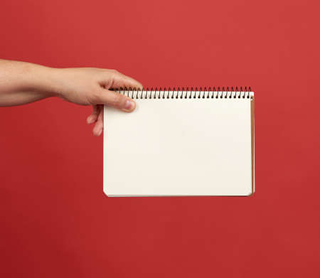 hand holding an open spiral notebook with empty white sheets, red background, copy space