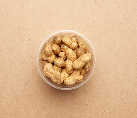 Inshell peanuts in a transparent plastic bowl on a brown wooden background, top view