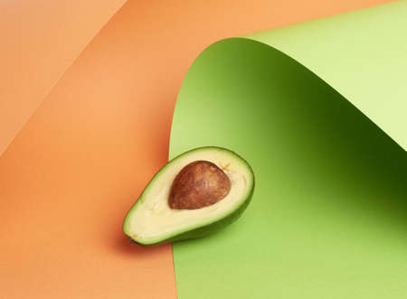 half ripe green avocado with a brown bone on an abstract green-orange background with curved paper