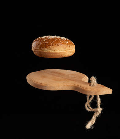 baked round bun with sesame seeds hover over brown wooden cutting board, black background, hamburger...