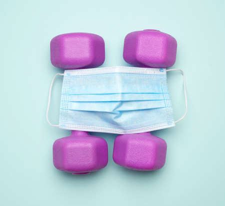 plastic dumbbells and medical mask. The concept of doing sports in protective equipment on a blue...