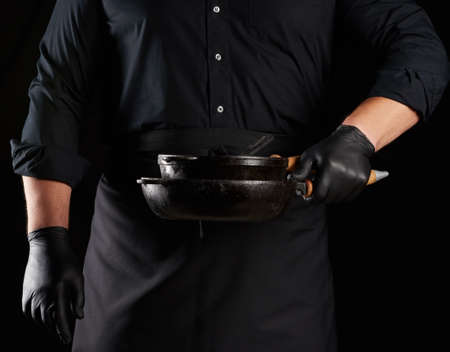 male cook in black uniform and latex gloves holds an empty round vintage black cast iron pan in front of him, low keyの写真素材