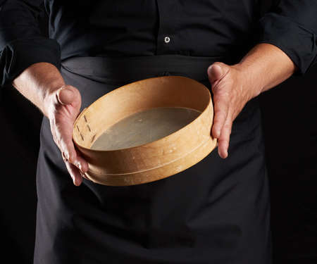 man in black uniform holding empty vintage round wooden sieve for sifting flour, chef stands against black background, close upの写真素材