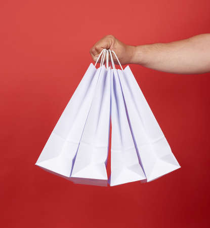 man in blue clothes holds a stack of white paper bags, red background, concept of shopping and...