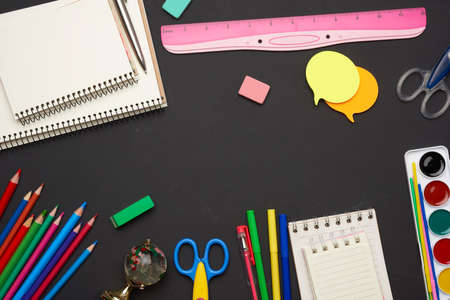 colorful wooden pencils, scissors, notepads and paint for drawing on a blank black chalk board,...