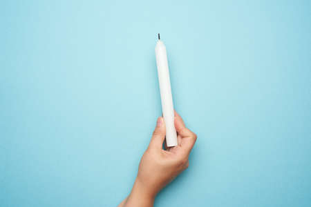 female hand holding a white wax candle on a blue background, copy spaceの写真素材