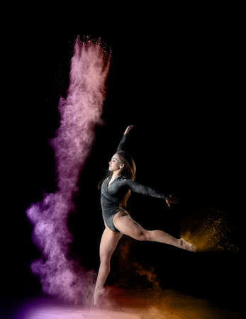 beautiful young caucasian woman in a black bodysuit with a sports figure dancing in a purple-yellow cloud of flour on a black background, explosion and expression in motionの写真素材