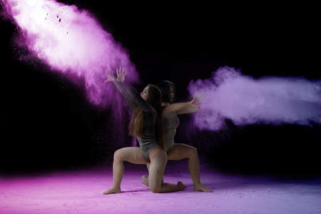 two beautiful young caucasian women in black bodysuits with a sports figure are dancing in a purple-blue cloud of flour on a black background, explosion and expresse in motionの写真素材