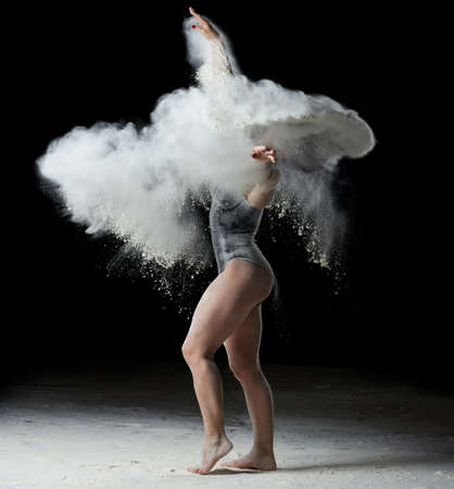 beautiful caucasian woman in a black bodysuit with a sports figure is dancing in a white cloud of flour on a black backgroundの写真素材