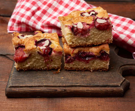 stack of square baked sponge cake slices with plums on wooden kitchen board, close upの写真素材